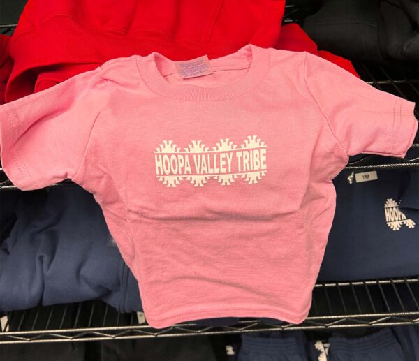 A pink baby tshirt sits on a rack, with the Hoopa Valley Tribe logo showing