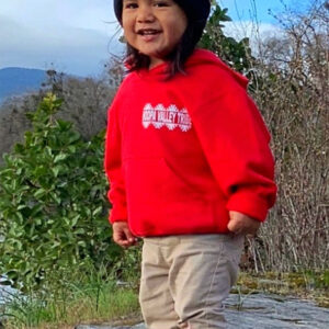 A smiling child is standing on a rock and wearing a red hooded jacket with the Hoopa Valley Tribe logo on it