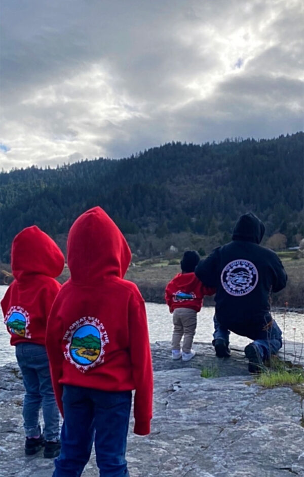 A family is standing on a rock overlooking a river and facing away from the camera. The three children are wearing red hooded jackets with the Hoopa Valley Tribe logo on them, and the adult is wearing a black hooded jacket with the Hoopa Valley Tribe logo on it.