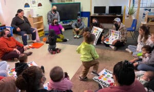 A number of adults and children sit in a classroom setting. Several of the adults are holding up posters, and the children are engaged in various activities.