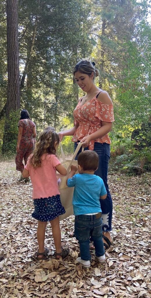 An adult stands in a forest speaking to two young children