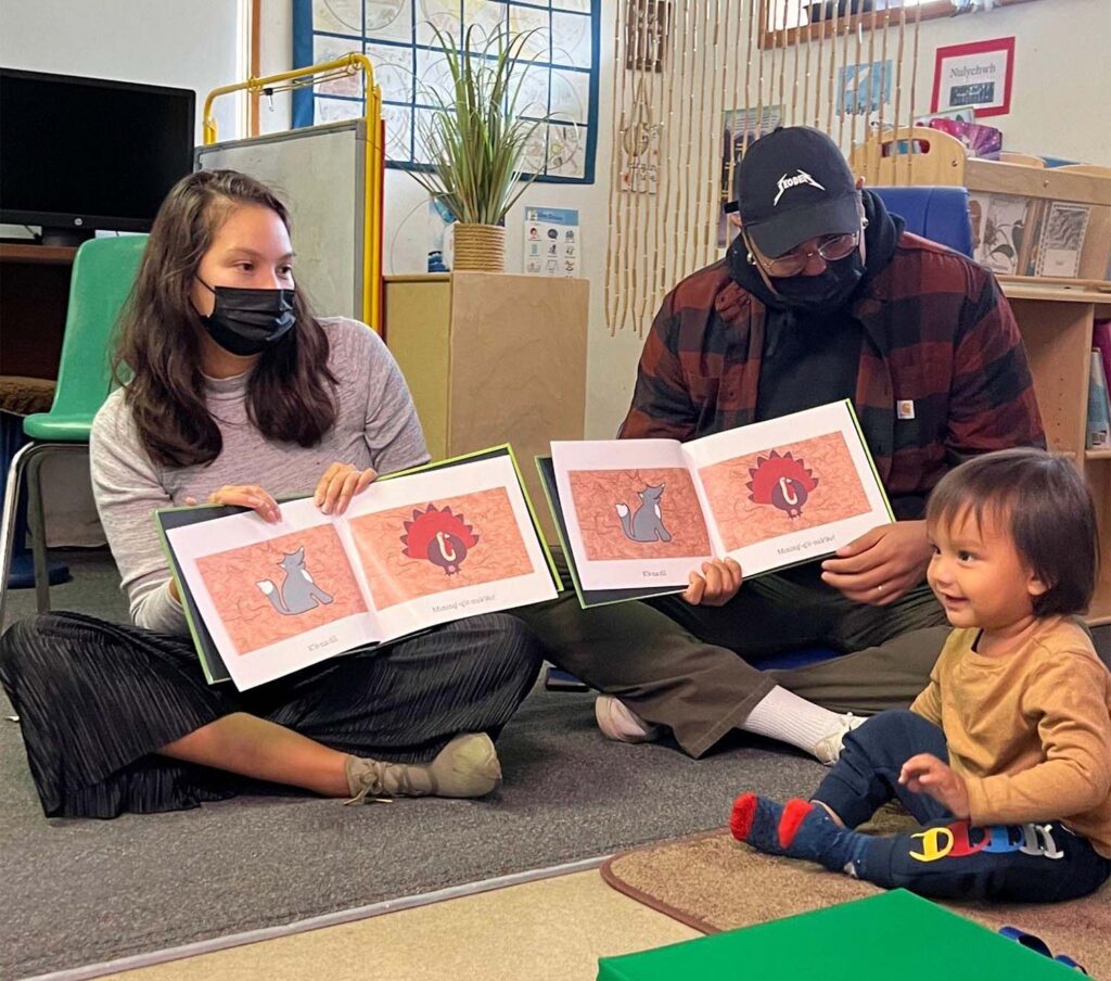Two adults sit in a classroom next to a young child. The adults are holding open a book for the child.