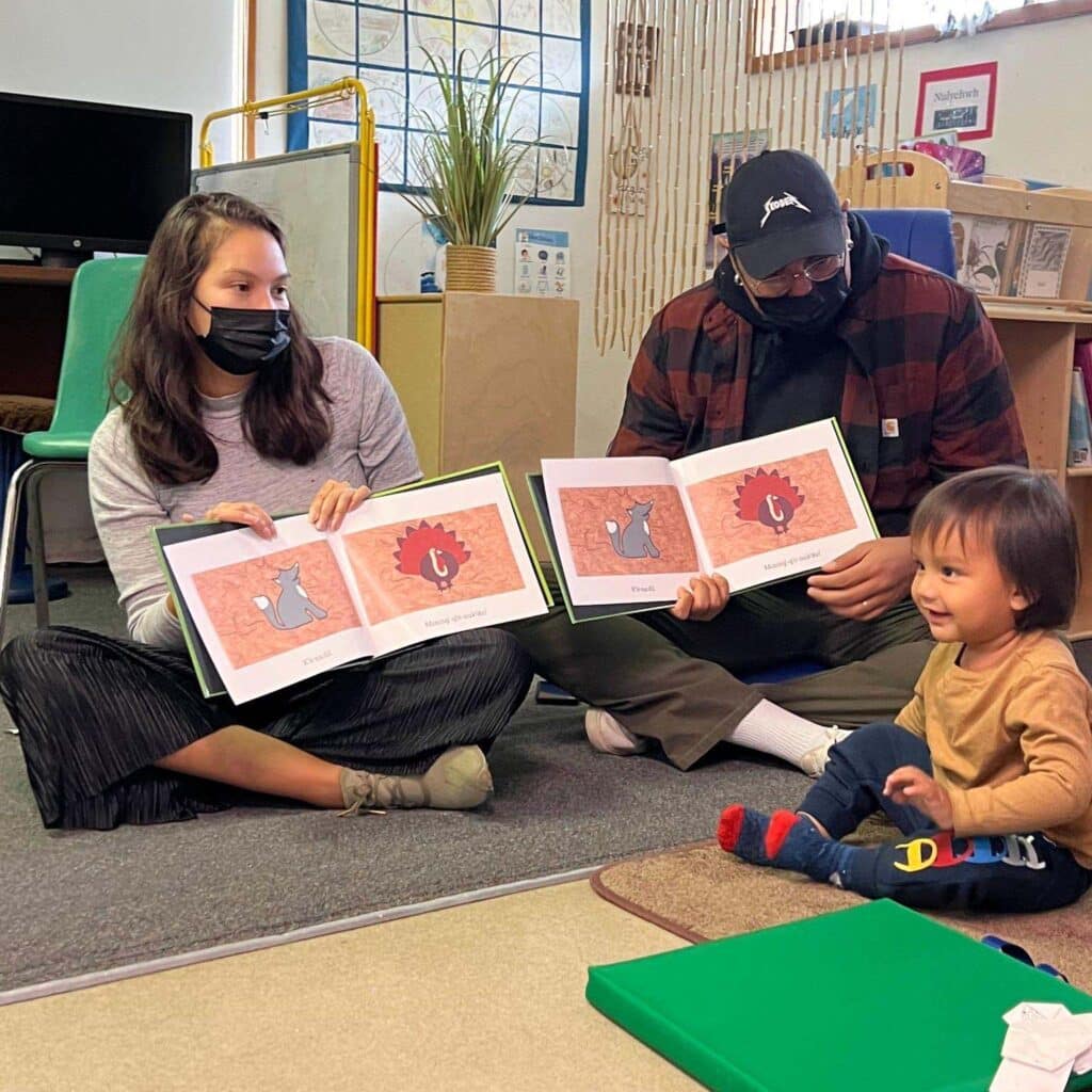 A family sits together on the floor in a classroom. The two parents are holding open picture books to the same page, while their young child smiles and crawls nearby.