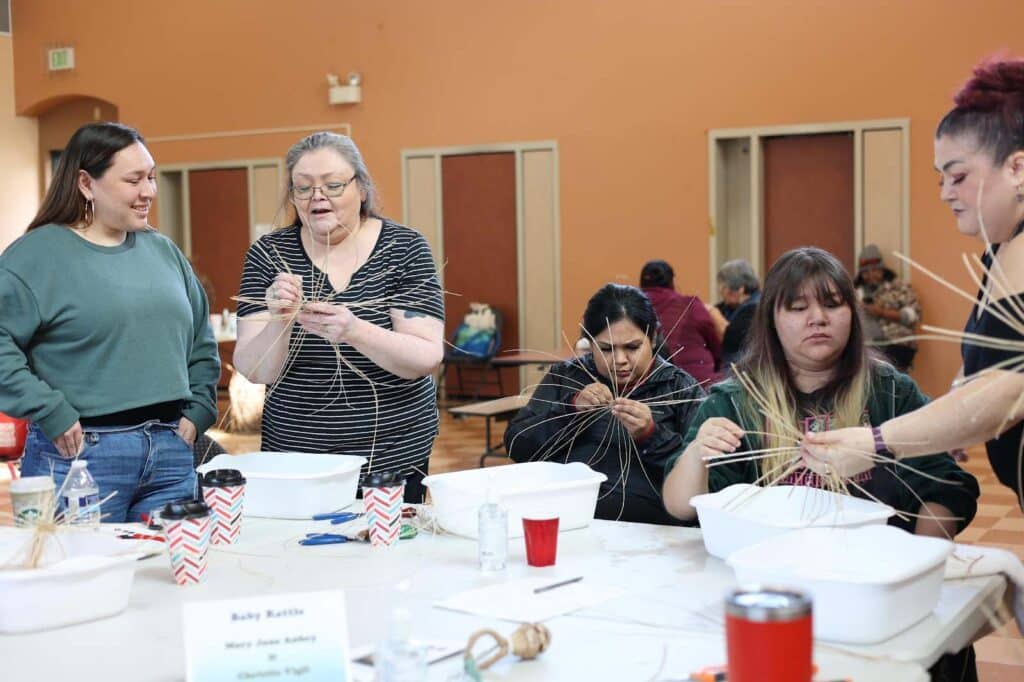 Several people sit around a table at various stages of weaving baskets. Some of the people are receiving instruction.