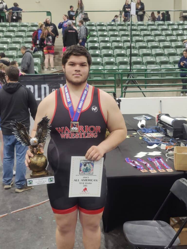 A youth wrestler in a gym holds a trophy and a certificate