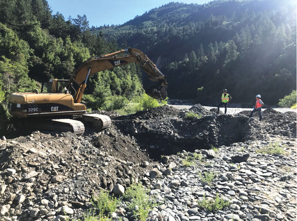 Some heavy equipment moves gravel on a river bed. Two workers in safety equipment stand on the river bed and watch